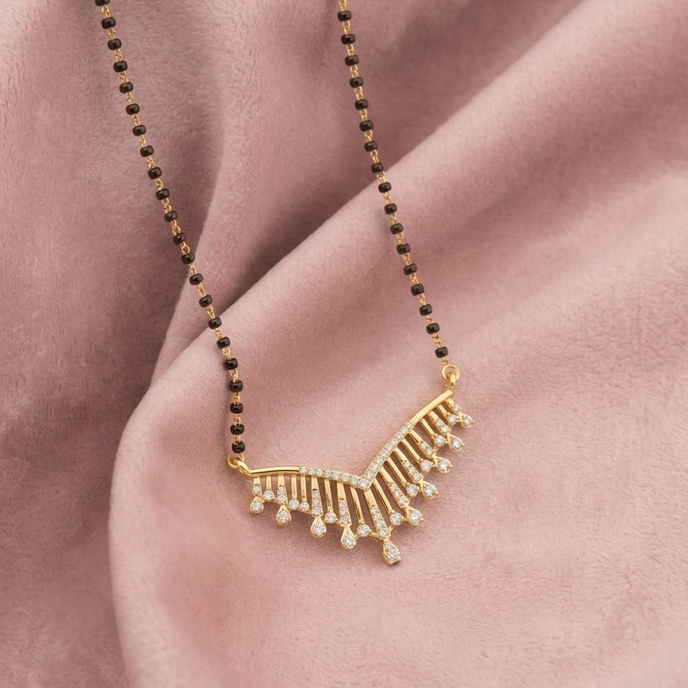 A yellow gold and diamond magalsutra with a curved, fan-like adorned with small clear stones and dangling teardrop charms, displayed on a soft pink fabric background.