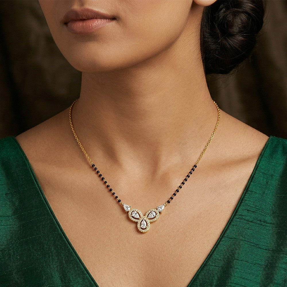 A close-up of a woman wearing a delicate gold mangalsutra with a black beaded chain and a central teardrop-shaped set with clear stones, paired with a dark green satin V-neck top.