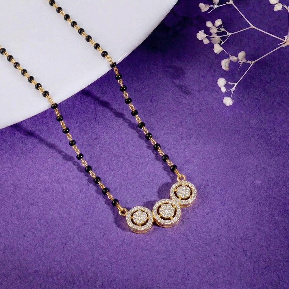 A yellow gold and diamond mangalsutra with three circular crystal-encrusted , displayed on a textured purple surface with a white curved prop and small white flowers.