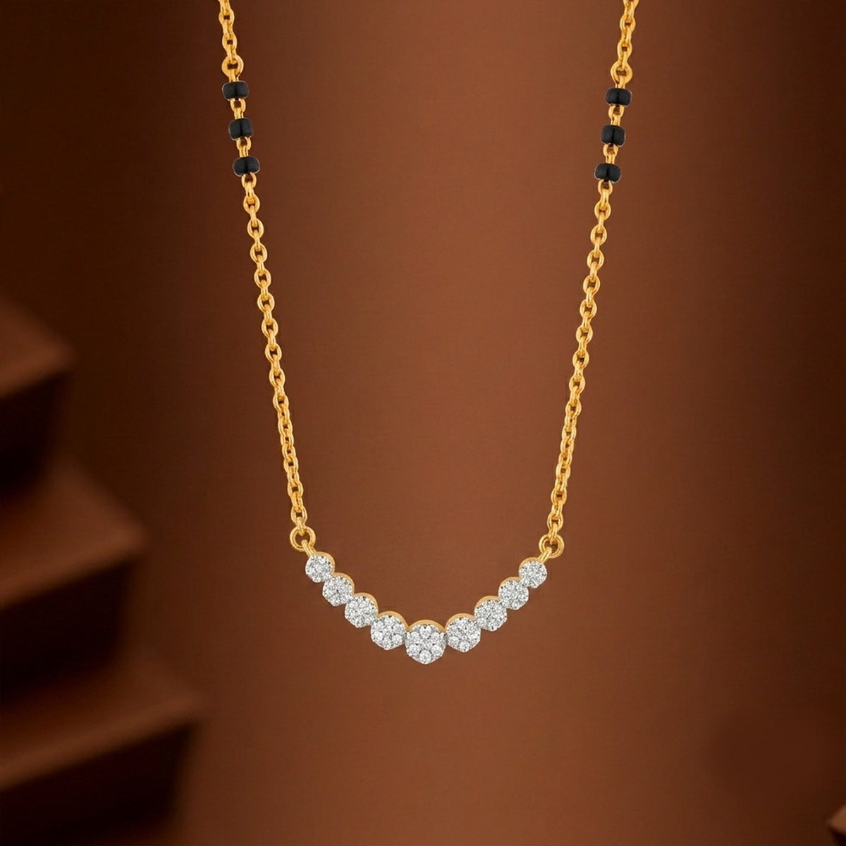 A gold chain mangalsutra featuring a curved row of clear pavé-set stones at the center, with small black bead accents on the chain near the top, shown against a warm brown gradient background.