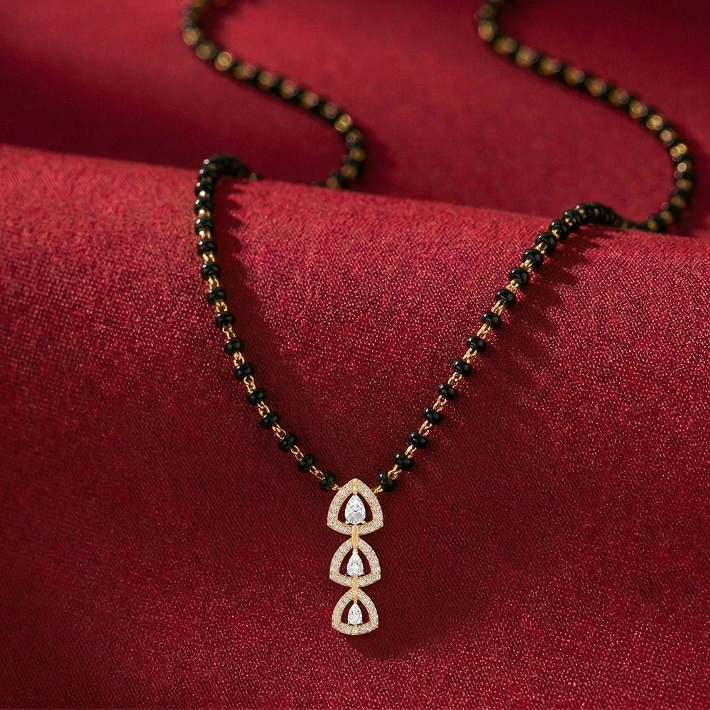 A black beaded mangalsutra with a gold-toned featuring three stacked teardrop motifs set with clear stones, displayed on a textured dark red fabric background.