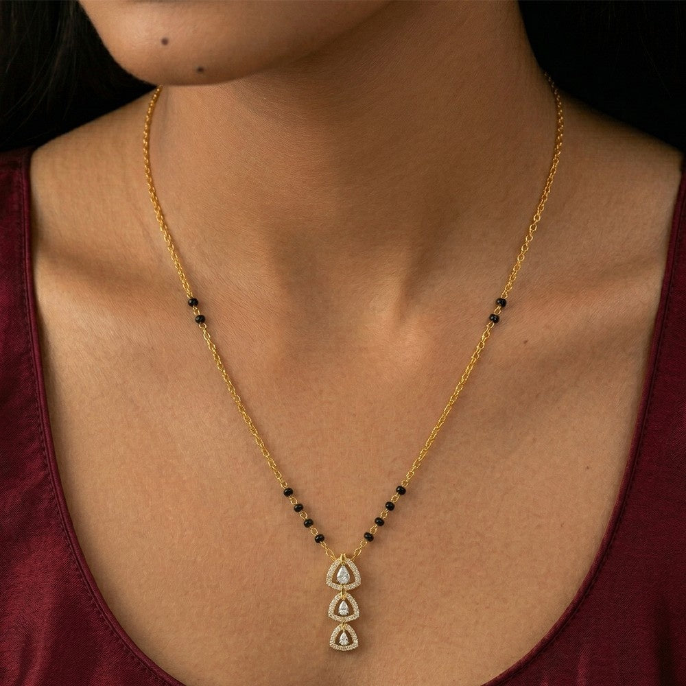 A close-up of a woman wearing a delicate yellow gold mangalsutra with small black beads and a teardrop-shaped with clear stones.