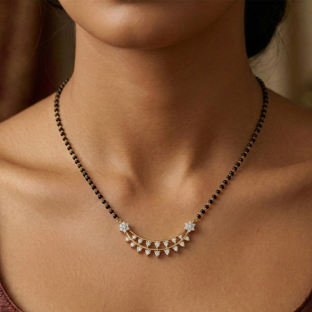 A close-up of a woman's neck and upper chest wearing a delicate mangalsutra. The mangalsutra has a black beaded upper section and a gold-toned lower crescent-shaped section adorned with small clear stones and star-like floral details.