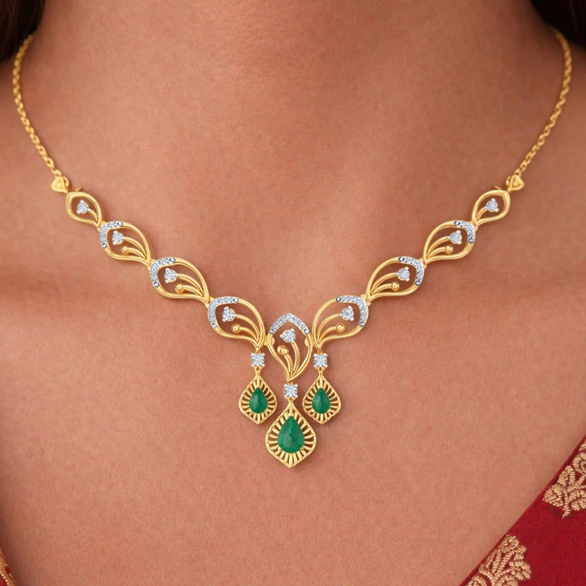 Gold necklace with a symmetrical leaf-shaped design, and two green teardrop gemstone, shown worn on a model's neck.