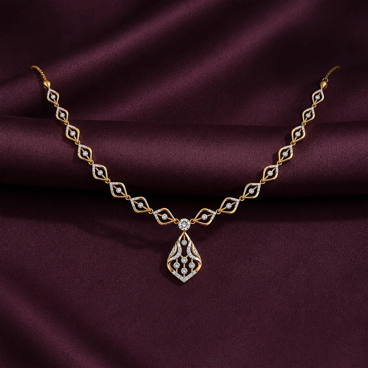 A gold necklace with a teardrop-shaped and repeating diamond motifs, displayed on draped dark burgundy satin fabric.