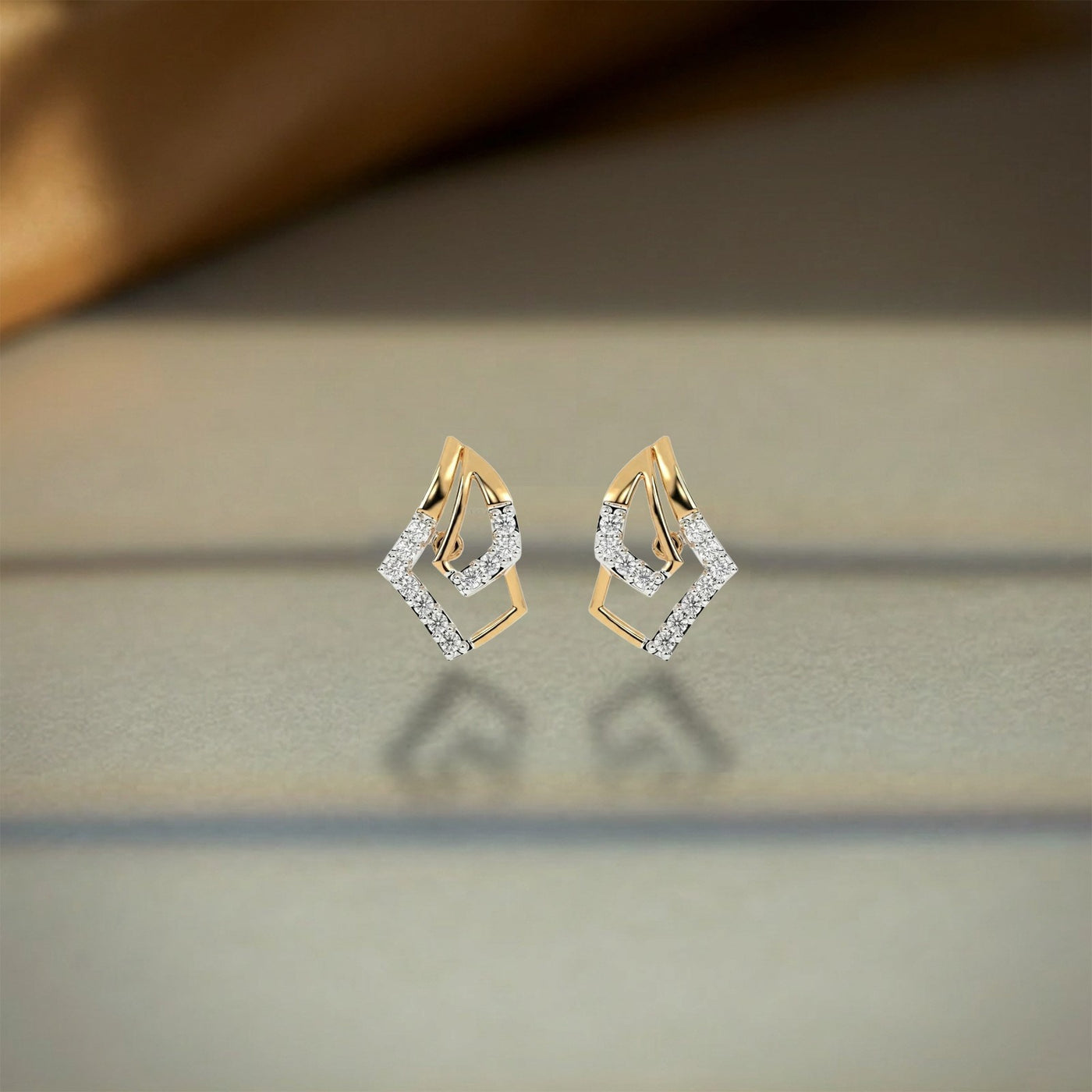 A pair of gold and diamond geometric stud earrings, displayed on a reflective surface with a soft-focus background.
