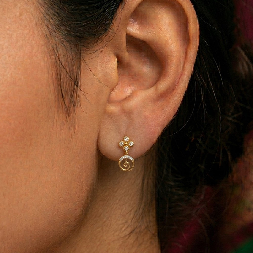 A close-up of a person's ear wearing a gold earring with a small floral top and a spiral drop.