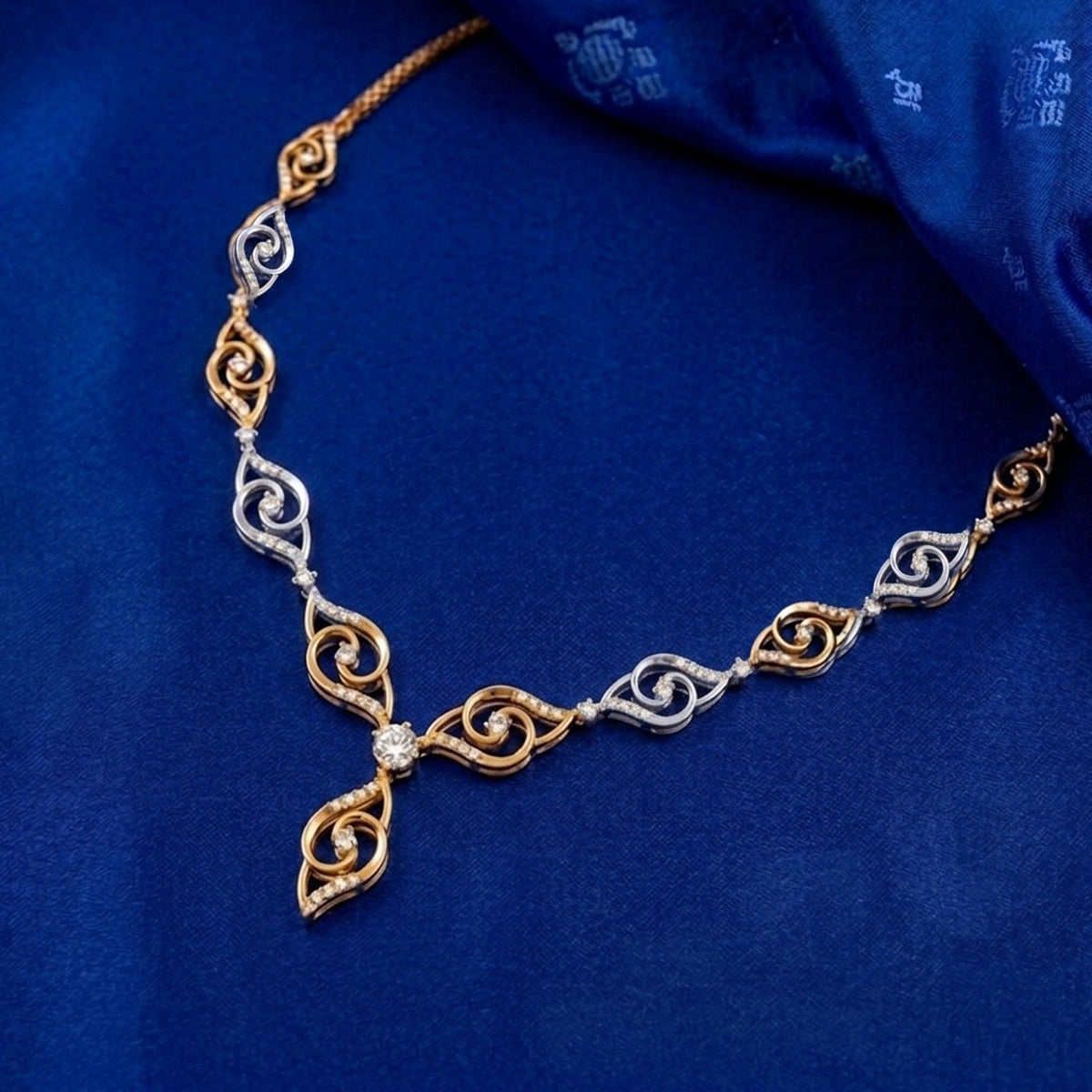 A delicate necklace with gold and dismond scrollwork links and a central diamond, displayed on a textured royal blue fabric background.