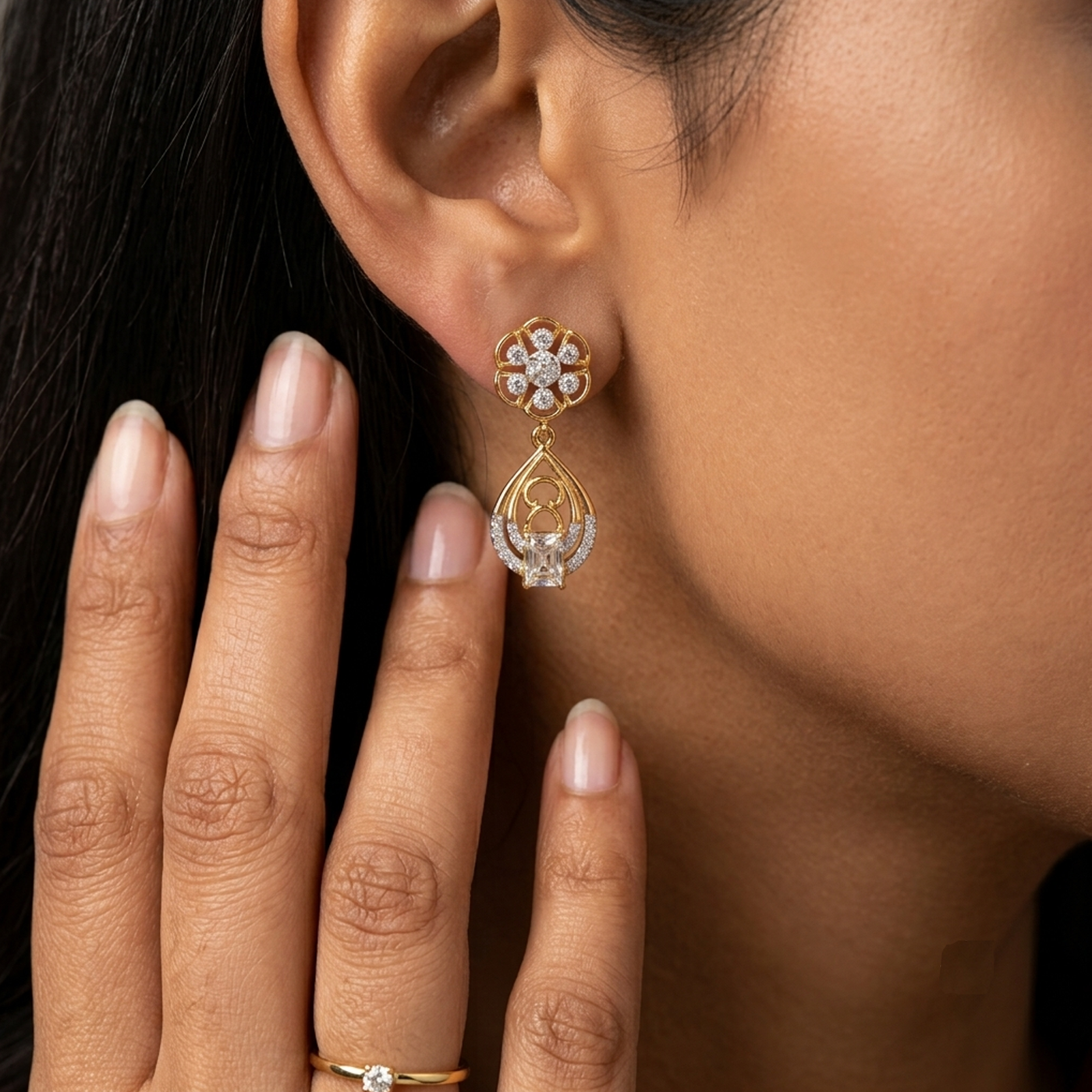 A close-up lifestyle shot of a woman wearing a gold and diamond earring. The earring features a floral stud with clear stones and a dangling teardrop-shaped gold frame encrusted with diamonds.