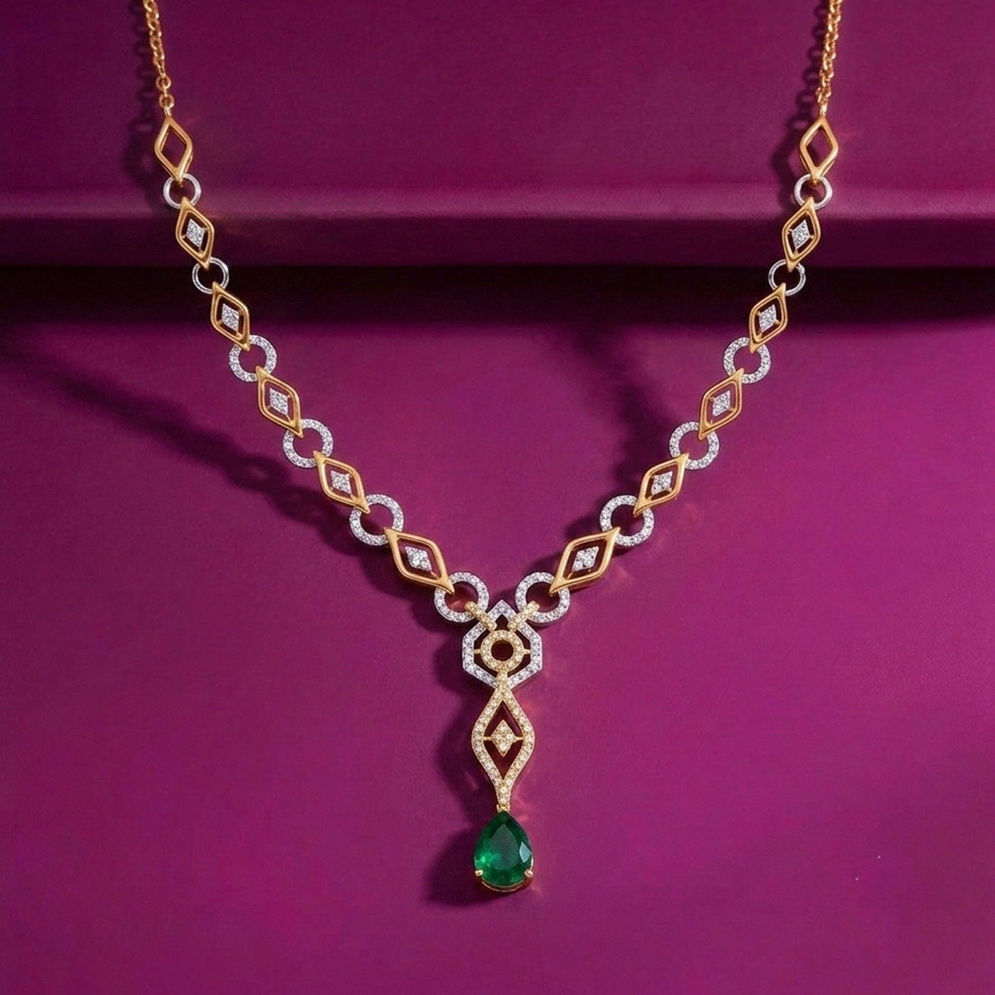 A gold and diamond geometric link necklace with a green teardrop emerald, displayed on a deep magenta background.