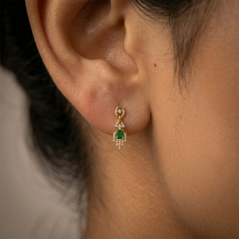 A close-up of a person's ear wearing a gold and diamond dangler earrings with rectangular green emerald-cut center stones.