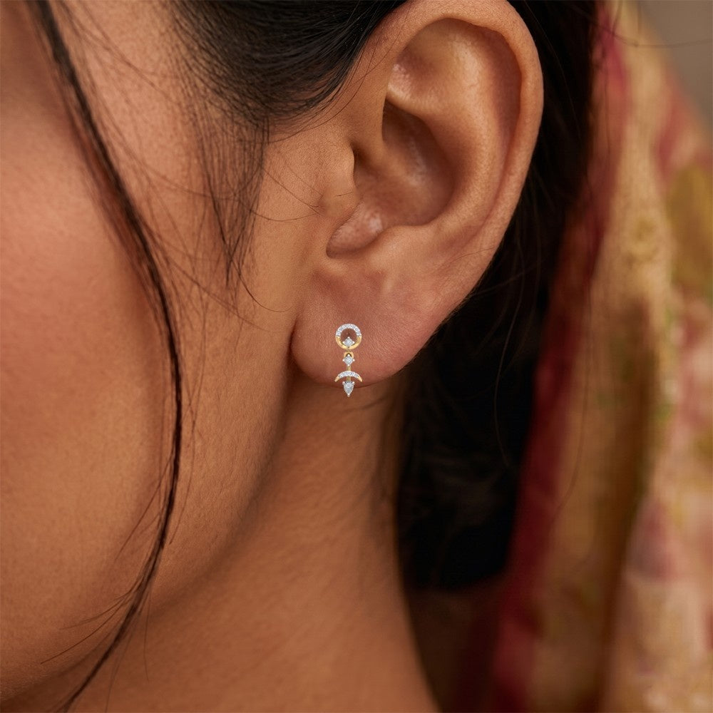 A close-up of a person's ear wearing a gold drop earring with multiple clear stones, set against a soft-focus background.