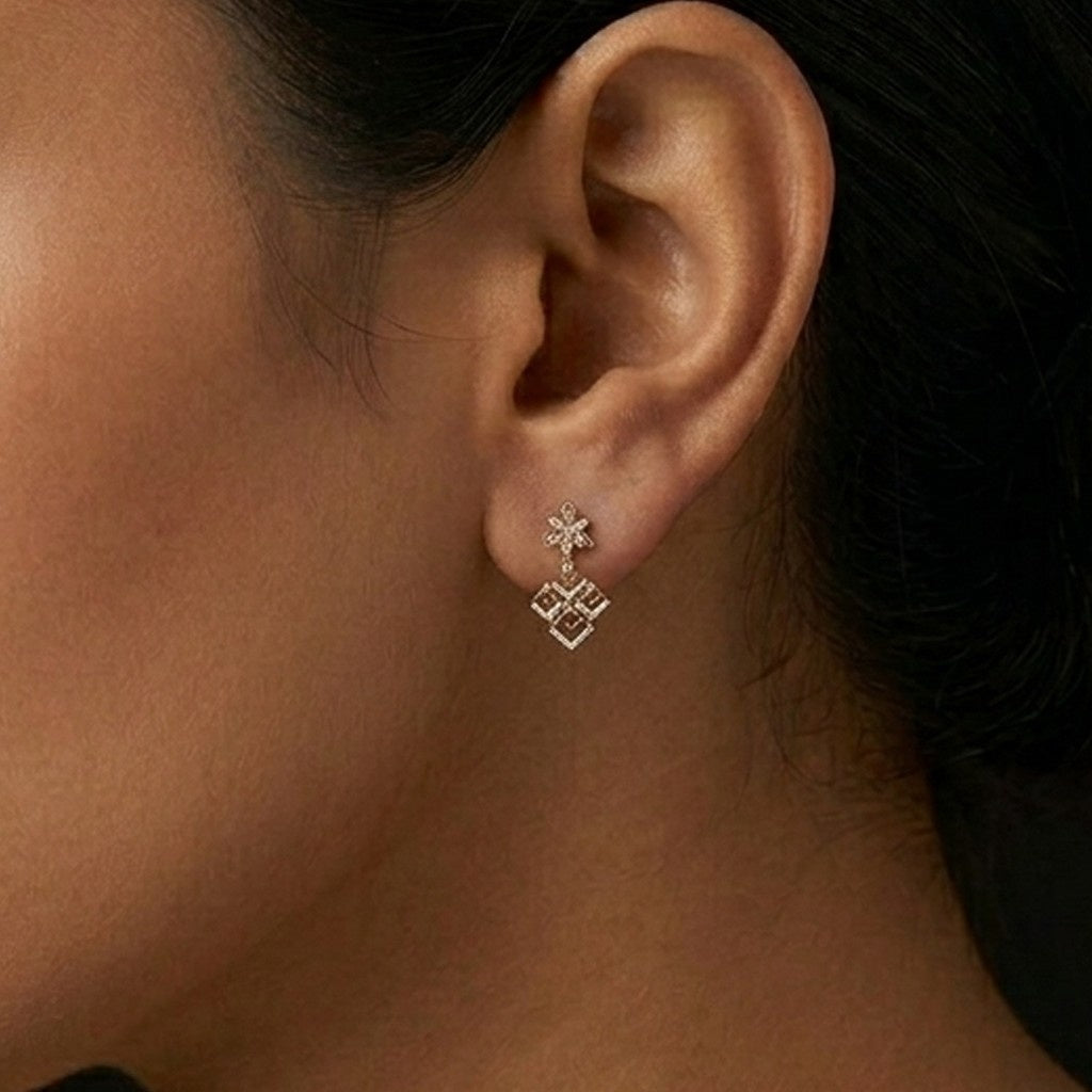 A close-up of a person's ear wearing a delicate gold dangle earring with a small floral stud and a geometric diamond-shaped dangle.