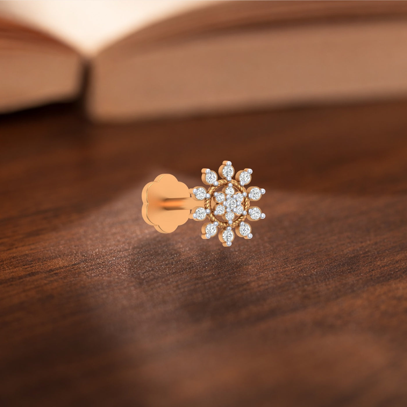 A single rose gold stud nose pin with a flower-shaped cluster of clear stones, shown on a dark wooden surface with a blurred book in the background.