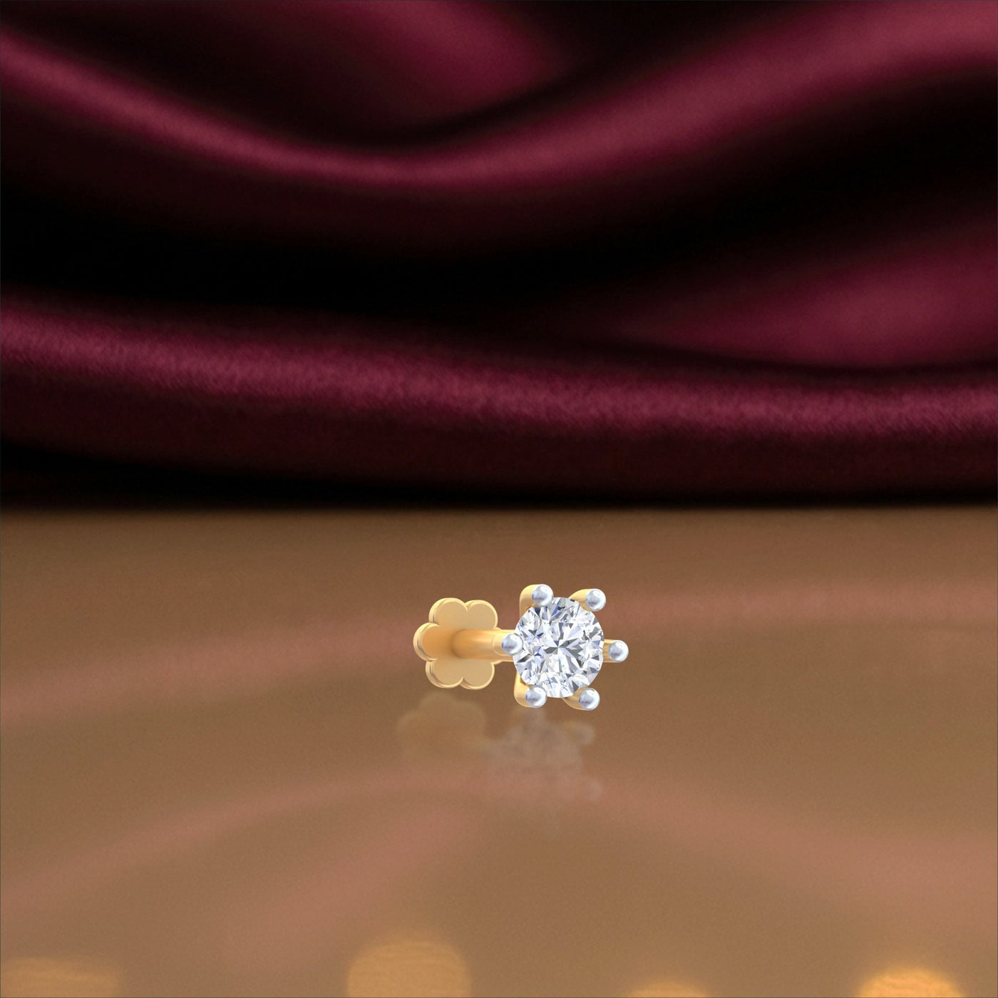 A single gold stud nose pin with a clear round-cut gemstone in a prong setting, shown on a reflective surface with a draped burgundy satin fabric background.