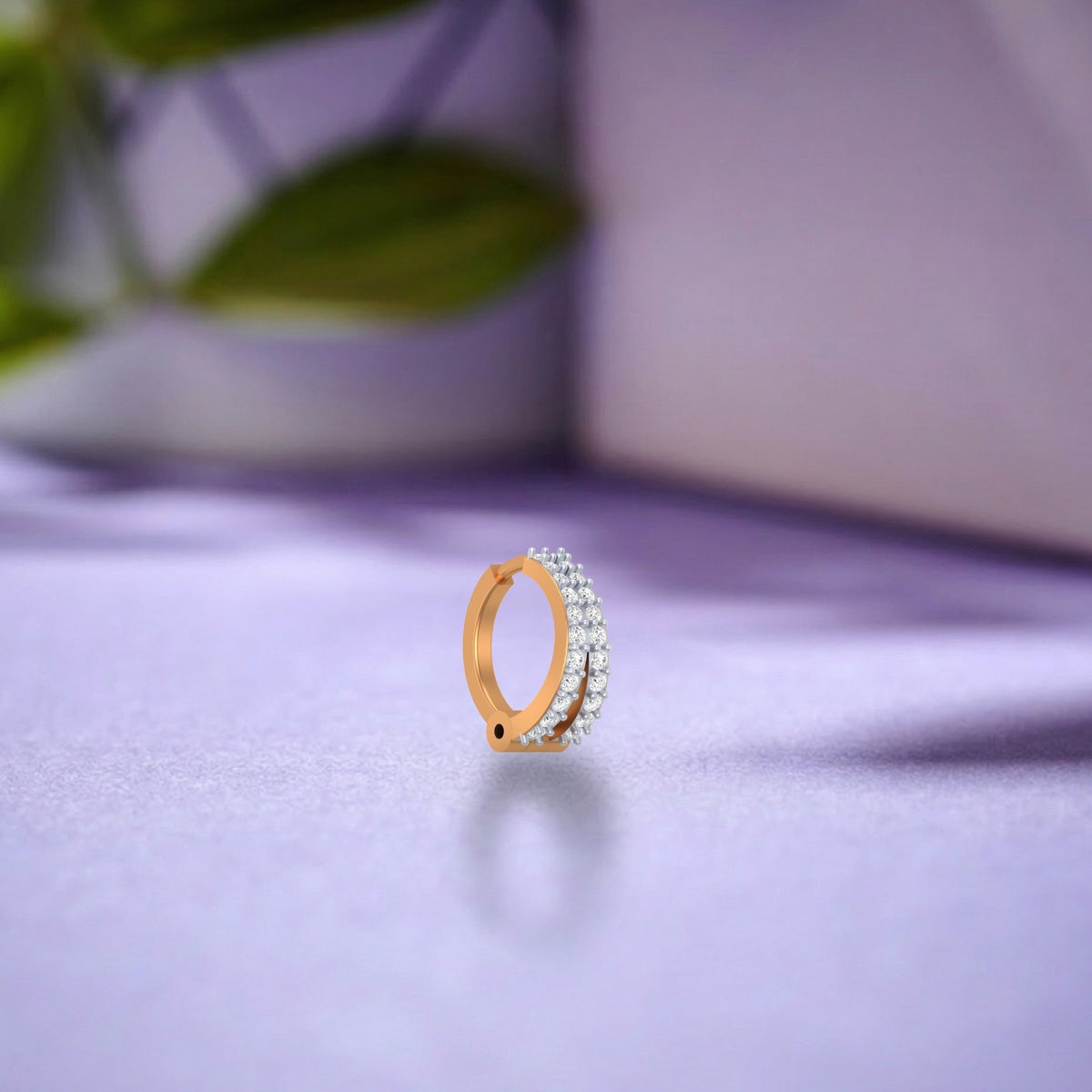 A small gold huggie hoop nose ring with a row of clear pavé stones on one half, standing upright on a soft lavender surface with a blurred background.