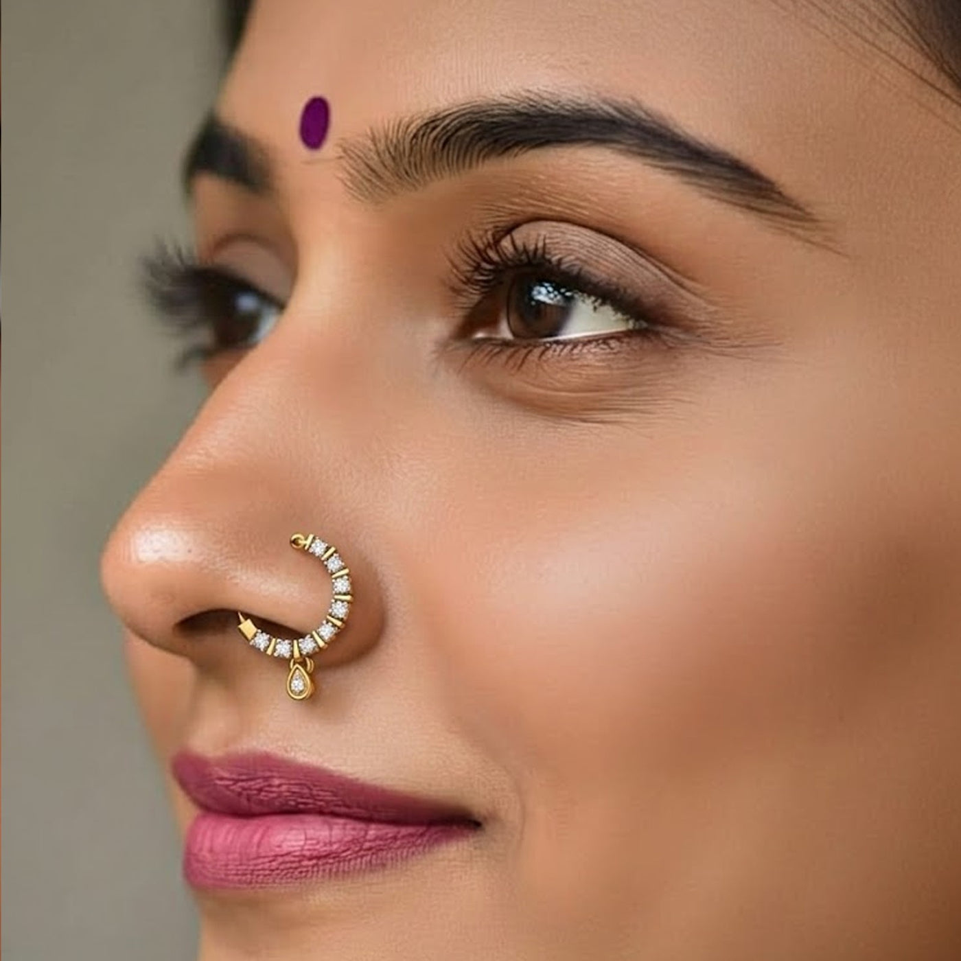 A close-up of a woman's face wearing a gold crescent-shaped nose ring adorned with small clear stones and a teardrop charm, with a purple bindi on her forehead.
