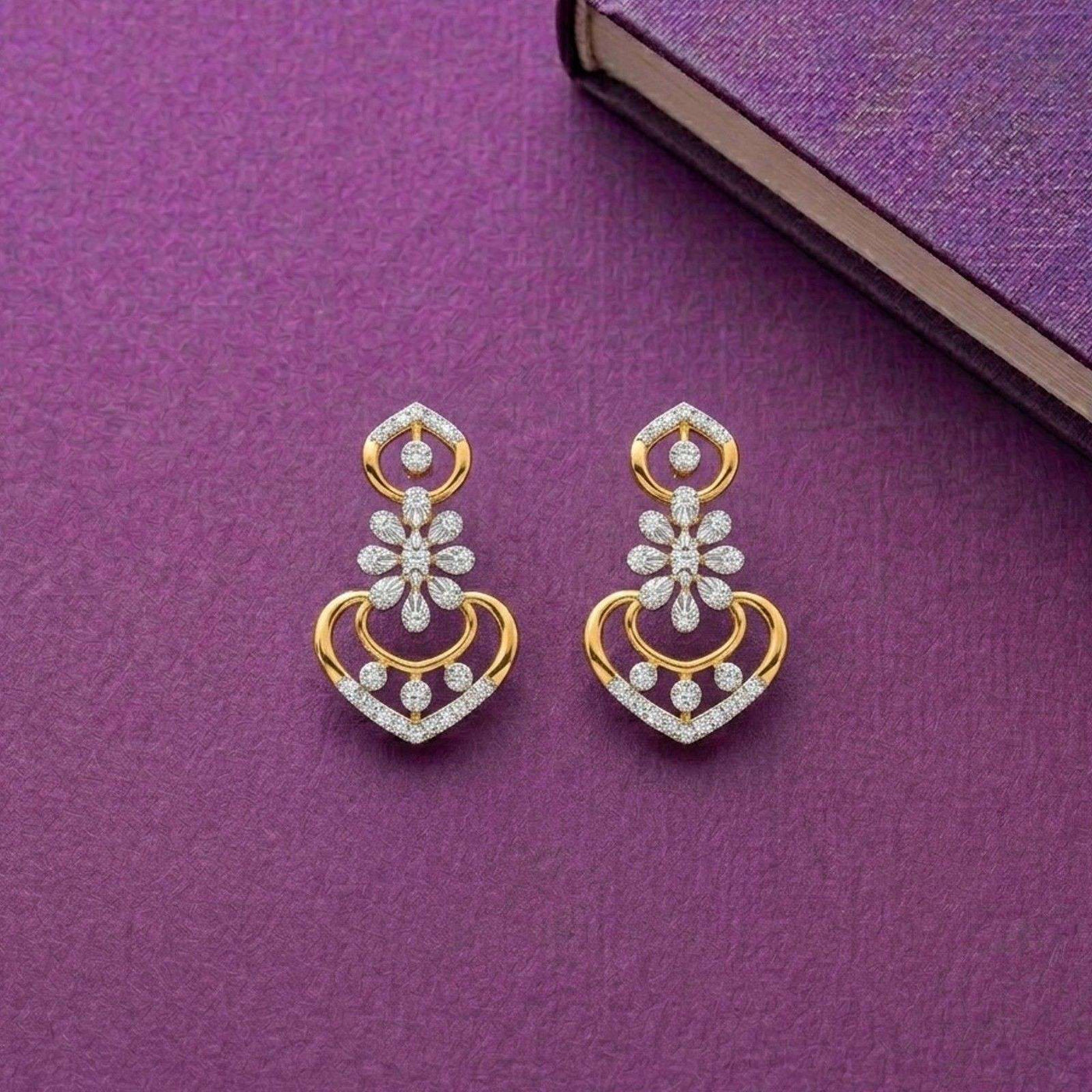 A pair of gold and diamond earrings, featuring a heart-shaped design with floral and circular details, displayed on a textured purple surface.