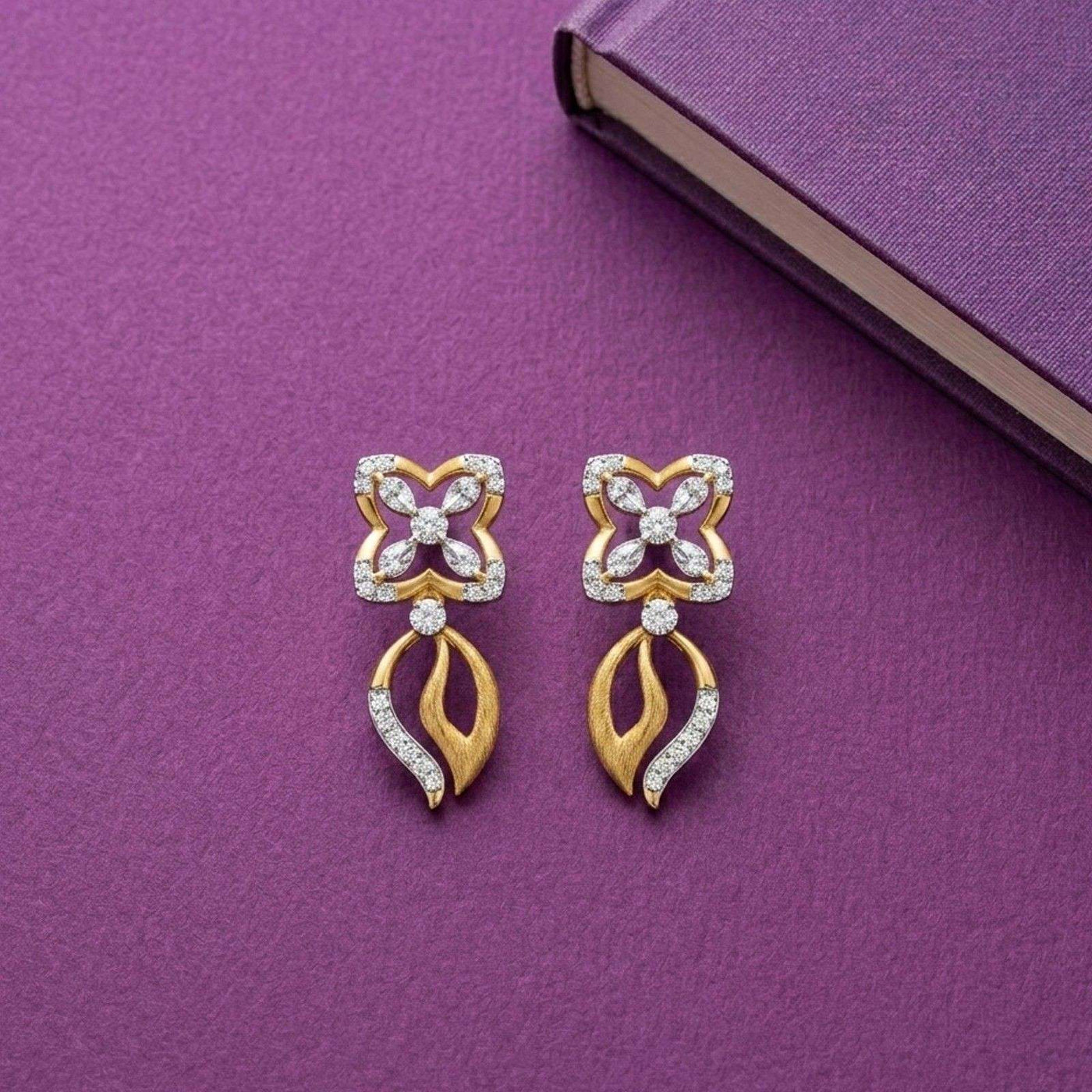 A pair of gold and diamond dangler earrings displayed on a textured purple surface next to a purple book.