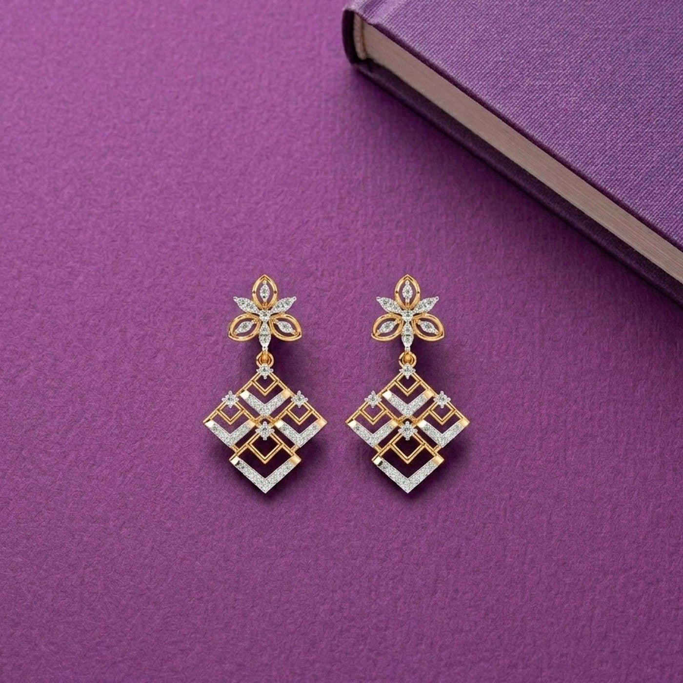 A pair of gold and diamond dangler earrings, featuring a floral stud top and a geometric diamond-shaped dangle on a purple surface.