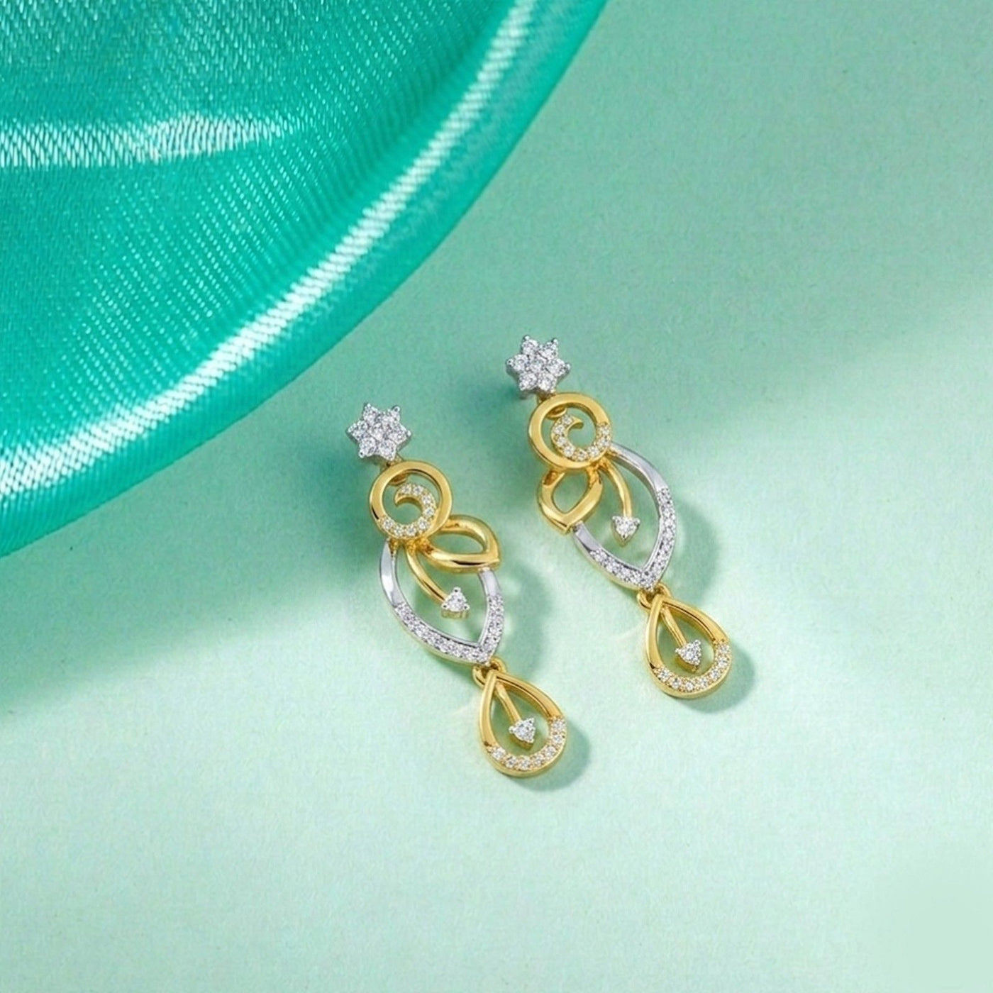 A pair of gold and diamond drop earrings, displayed on a light mint green surface.