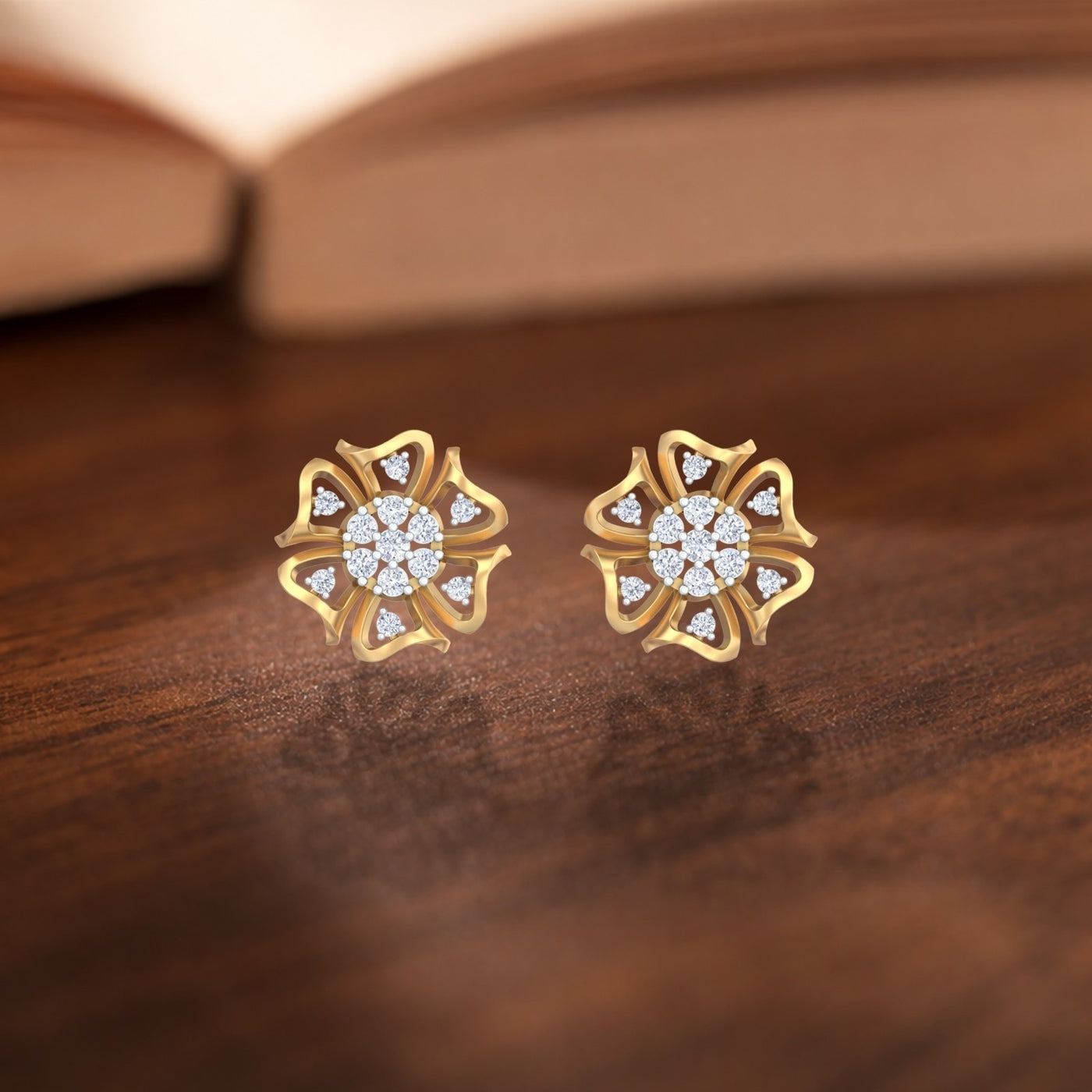 A pair of gold flower-shaped stud earrings, displayed on a dark wooden surface with a blurred book in the background.