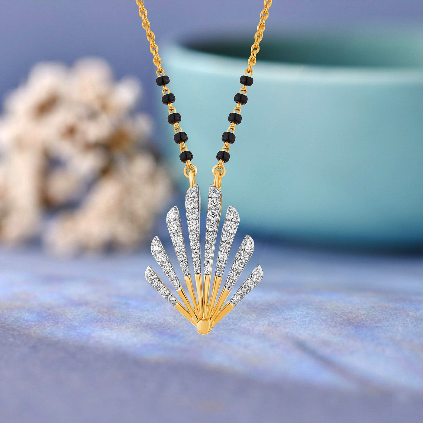 Gold fan-shaped mangalsutra with clear stones and a black beaded chain, displayed against a soft blue background with blurred decorative elements.