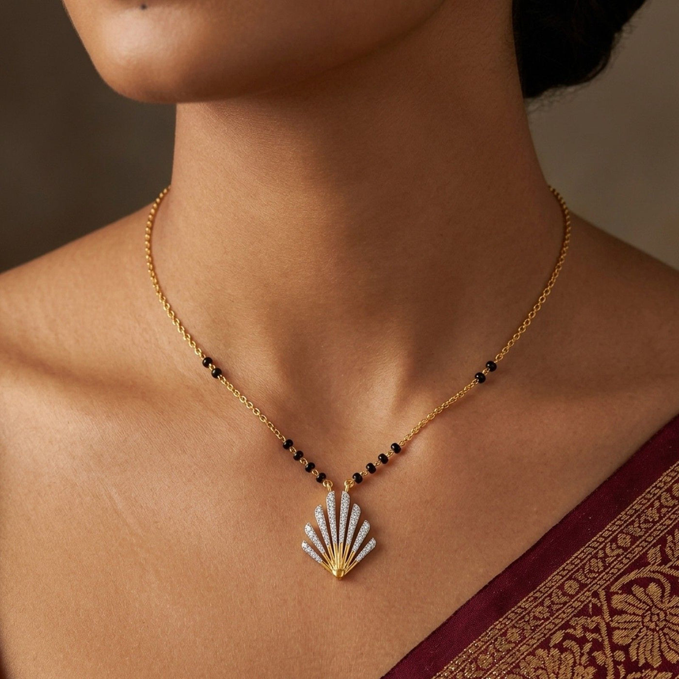 A close-up of a woman wearing a delicate gold mangalsutra with a shell-shaped mangalsutra. The pendant is set with white stones on one side and gold on the other, and the chain has small black bead accents.