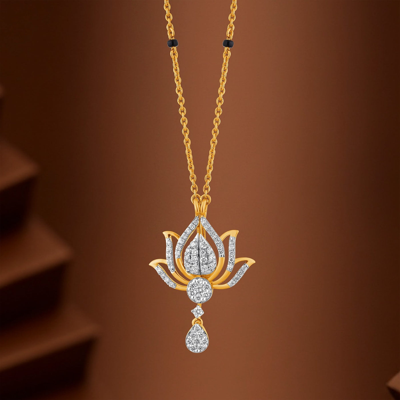 A gold mangalsutra featuring a lotus flower design adorned with clear stones and a teardrop-shaped dangling pendant, shown against a warm brown background.