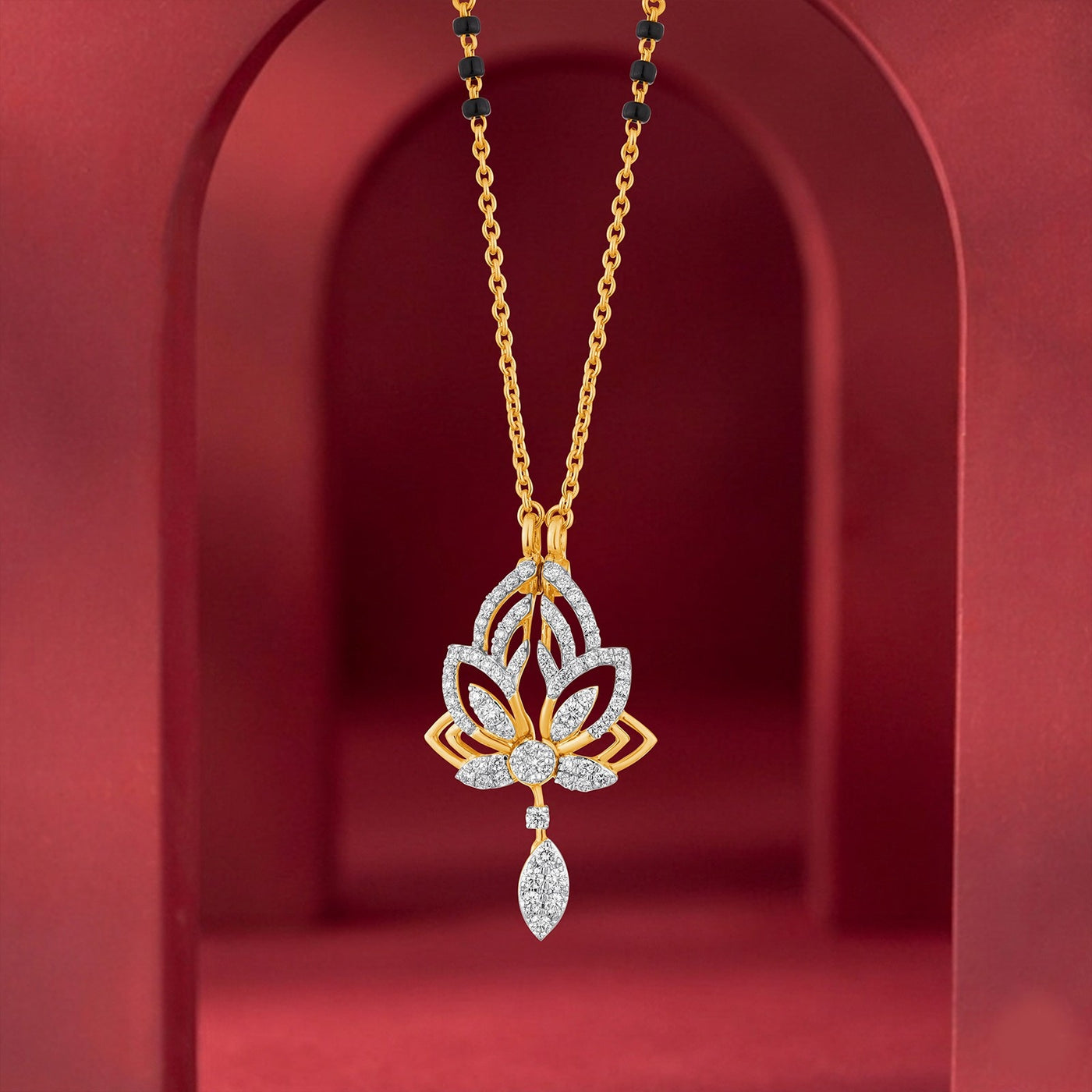 A gold and diamond mangalsutra featuring a lotus flower design with a teardrop-shaped diamond drop, displayed against a red arched background.