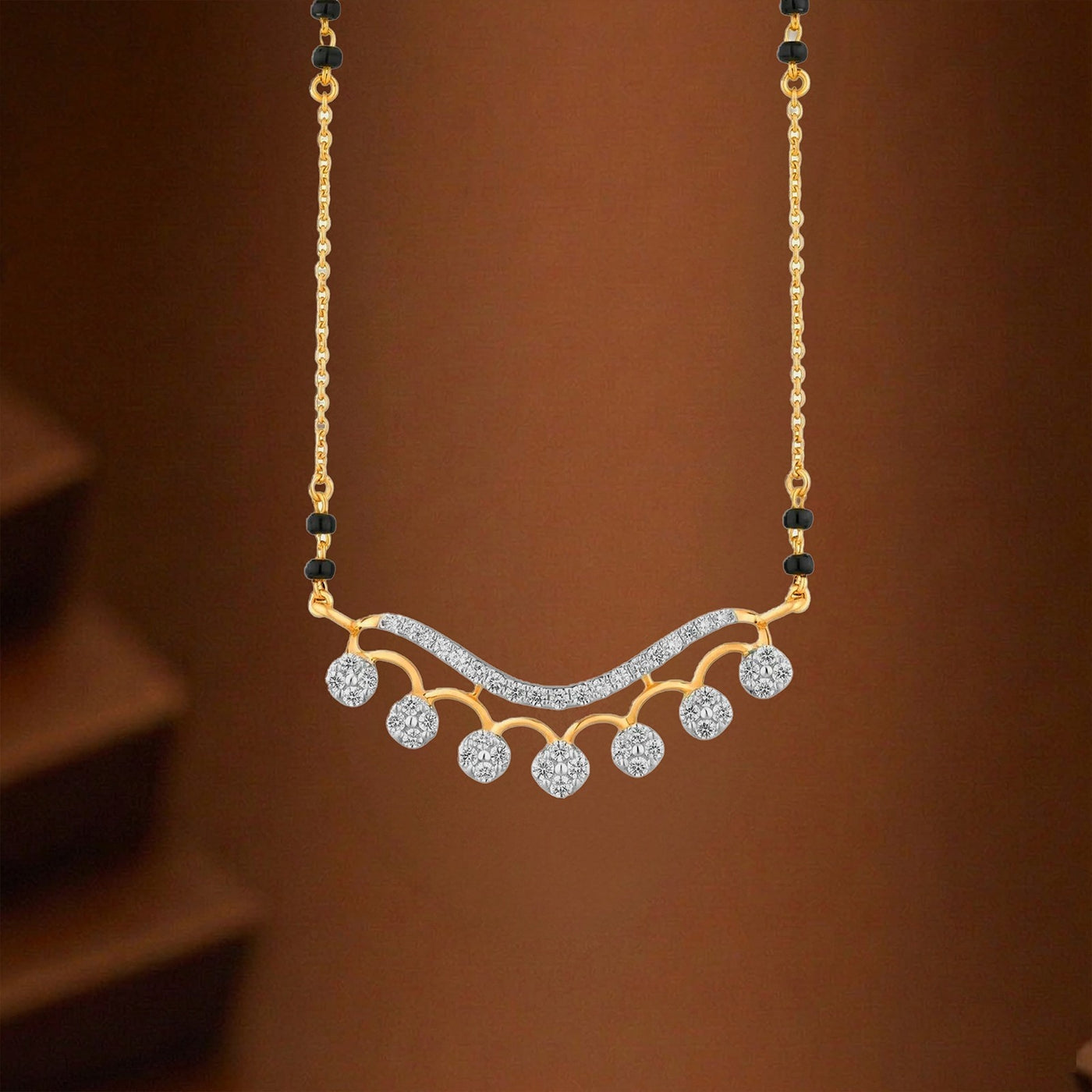 A gold mangalsutra with a wavy chain-link design, featuring five small round clear stones and a central curved row of clear pavé stones, suspended against a warm brown background.