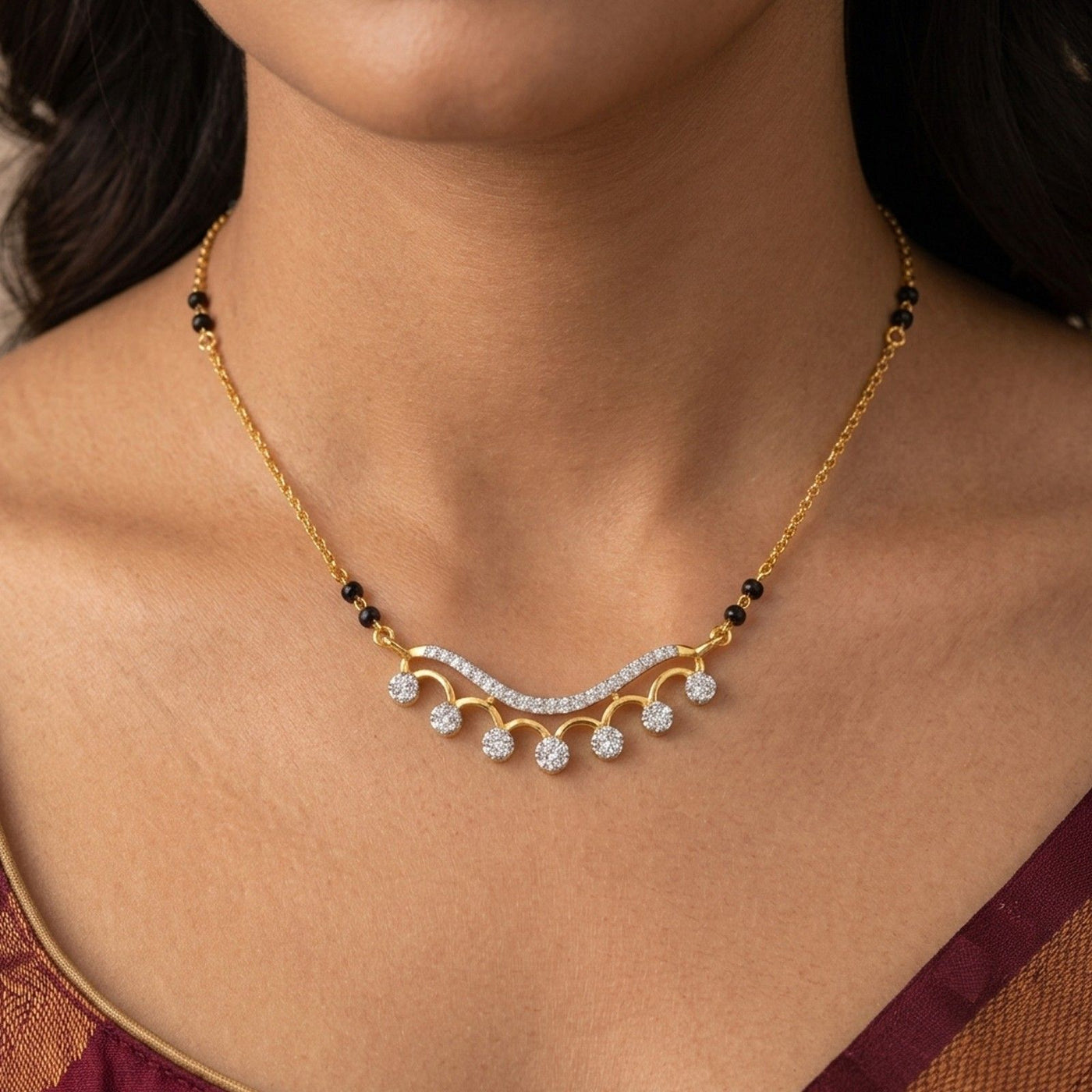 A close-up of a woman wearing a gold mangalsutra with a curved pavé-style center, small black beads, and several clear gemstone drops. 