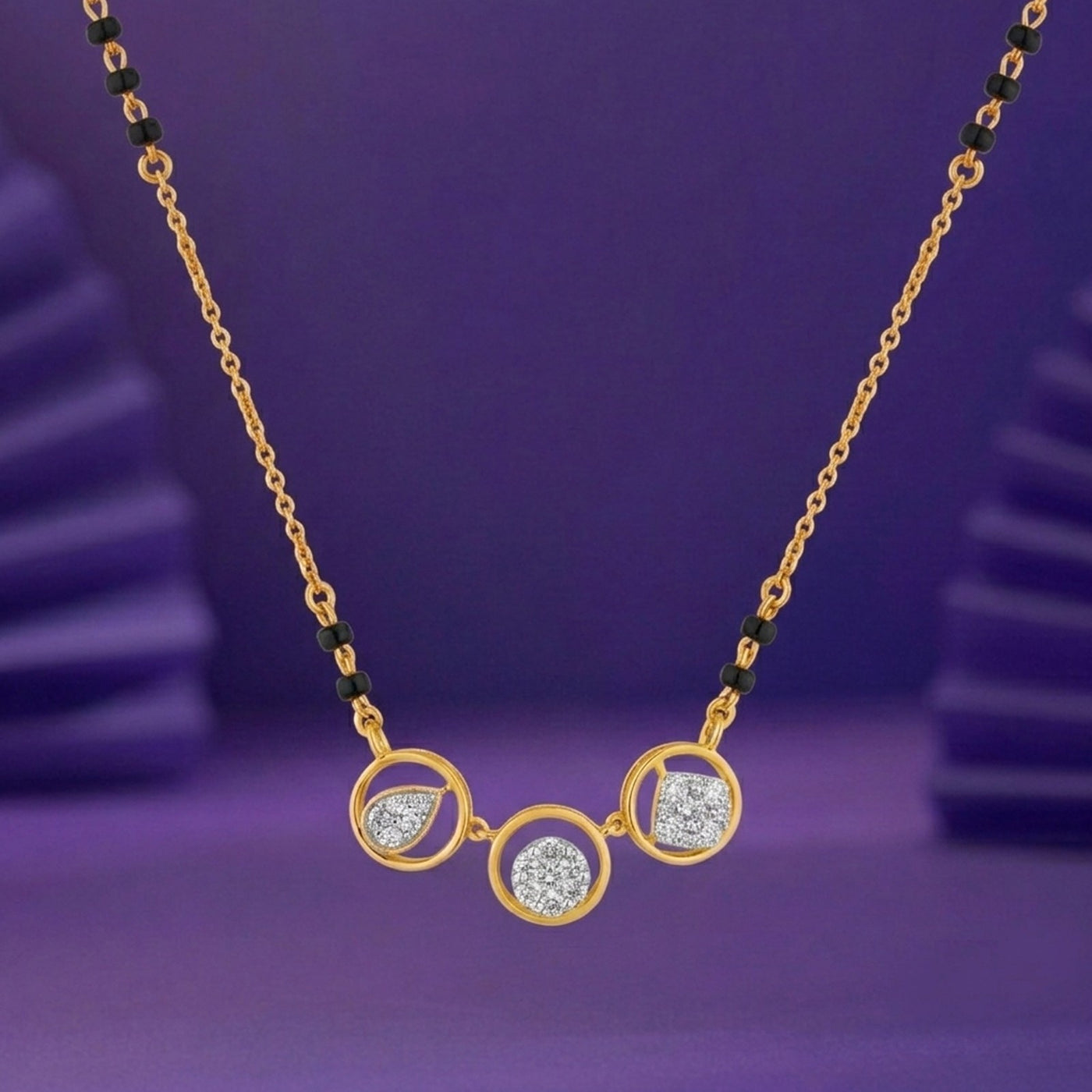A gold mangalsutra featuring three circular and teardrop-shaped settings, two of which are adorned with clear stones, set against a purple background.