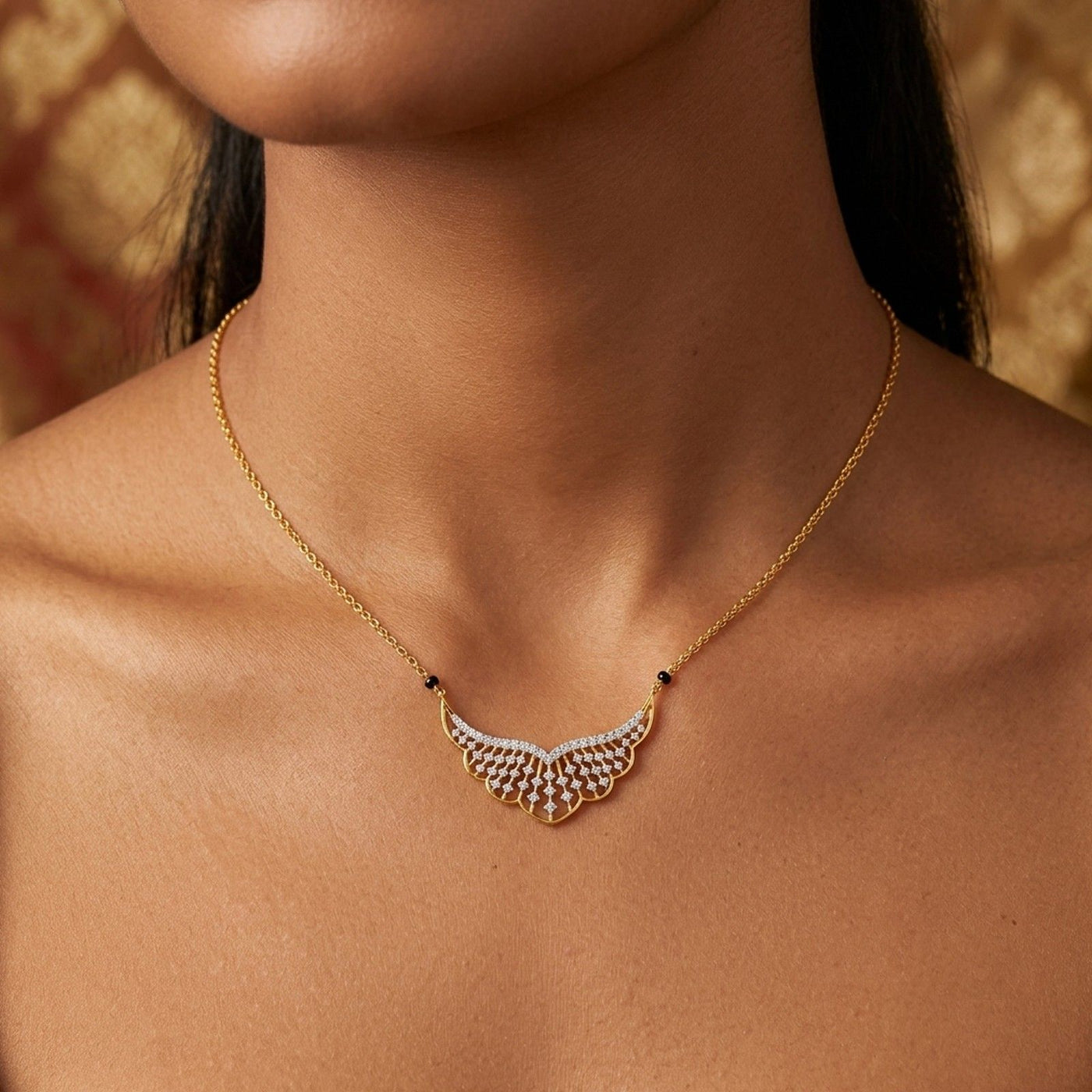 A gold mangalsutra with a wing-shaped with small clear stones, worn on a model's neck against a warm, patterned background.