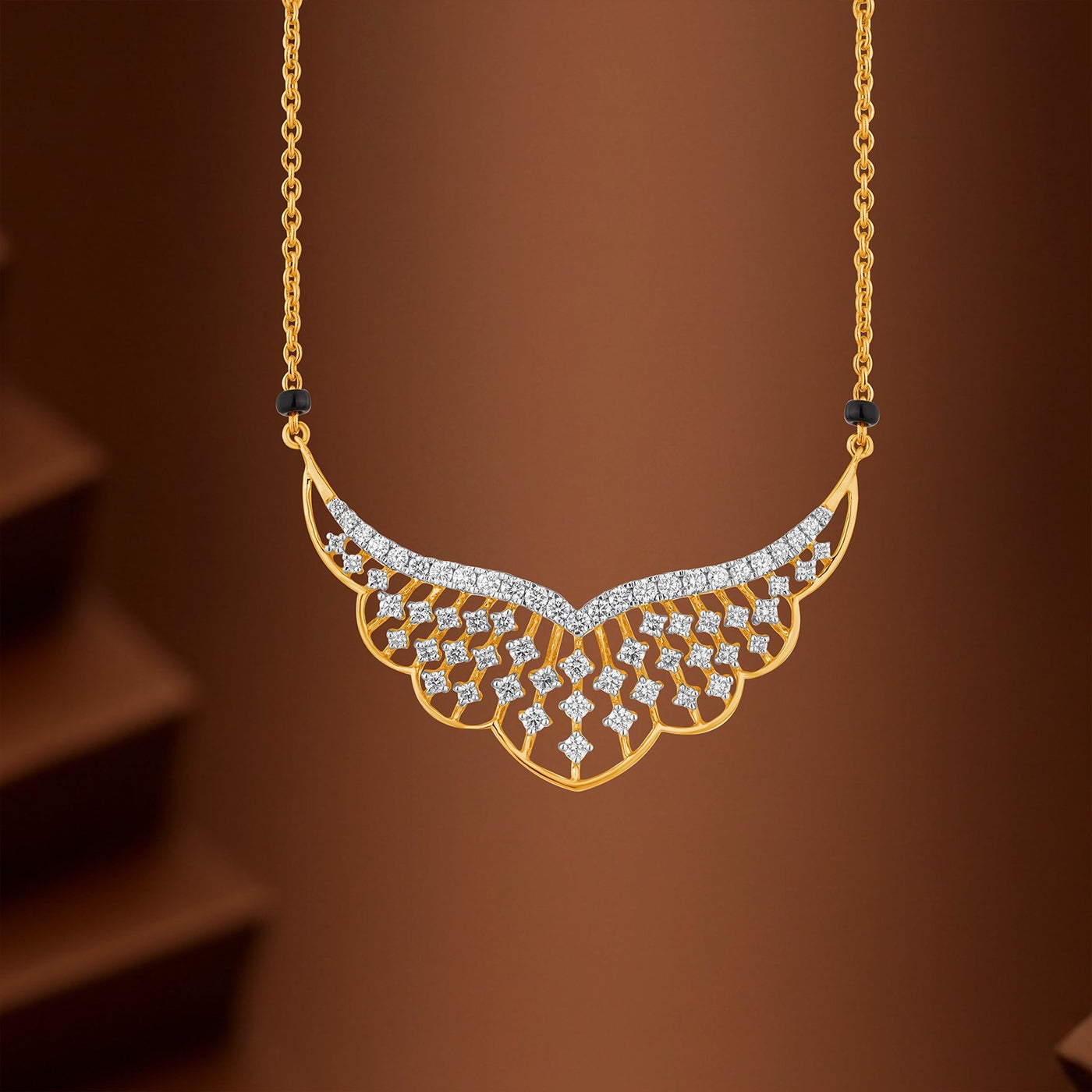 Gold wing-shaped mangalsutra with sparkling clear stones, suspended from a delicate gold chain against a warm brown gradient background.