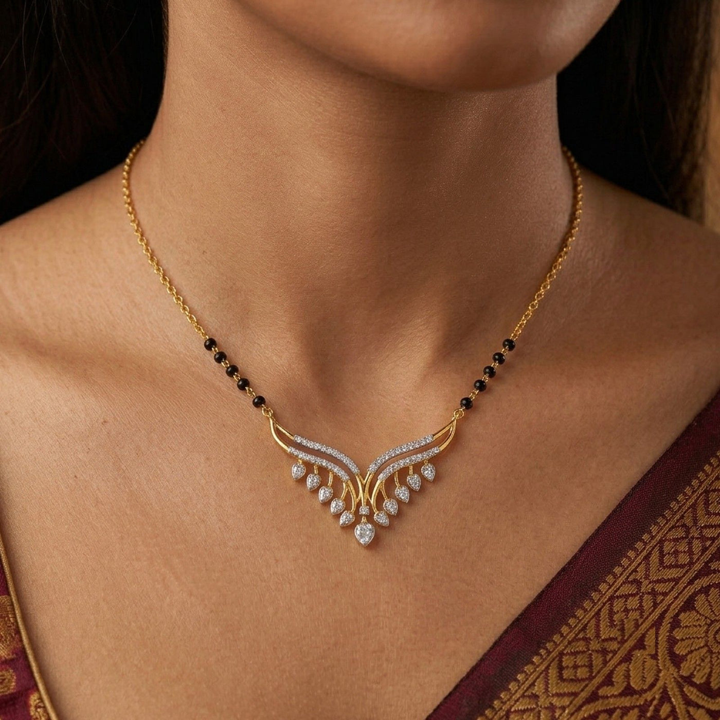 A close-up of a woman wearing a gold V-shaped mangalsutra with small black beads on the chain and clear stones forming a wing-like pendant.