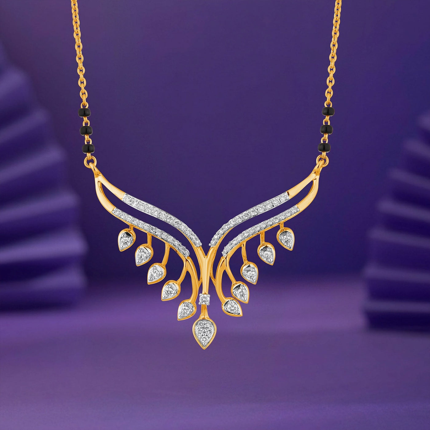 Gold wing-shaped mangalsutra with pavé-set clear stones and dangling teardrop-shaped stones, shown against a purple studio background.