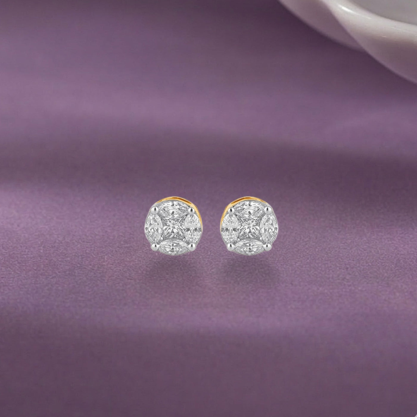 A pair of gold and diamond round stud earrings, displayed on a purple satin fabric background.
