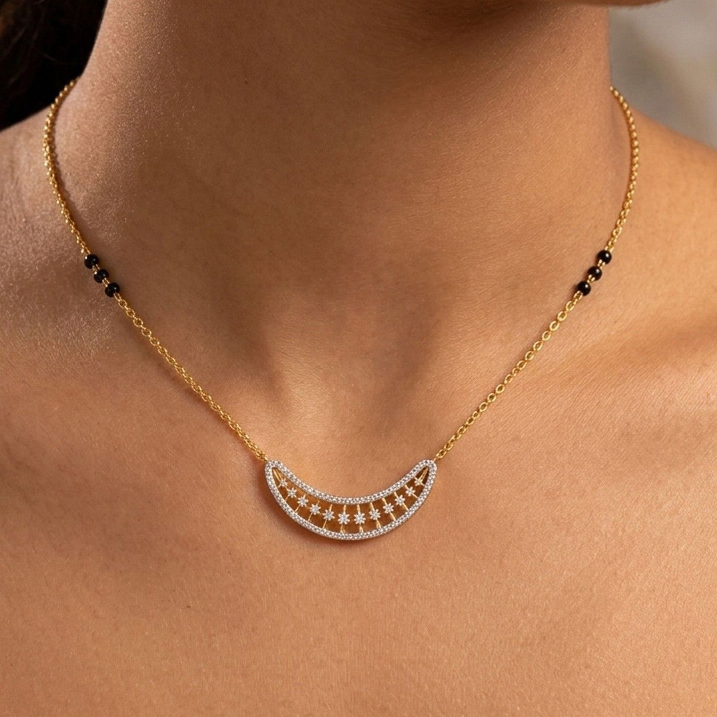 A close-up of a gold chain mangalsutra worn on a model's neck, featuring a crescent moon-shaped mangalsutra with a row of small clear stones and black bead accents on the chain.