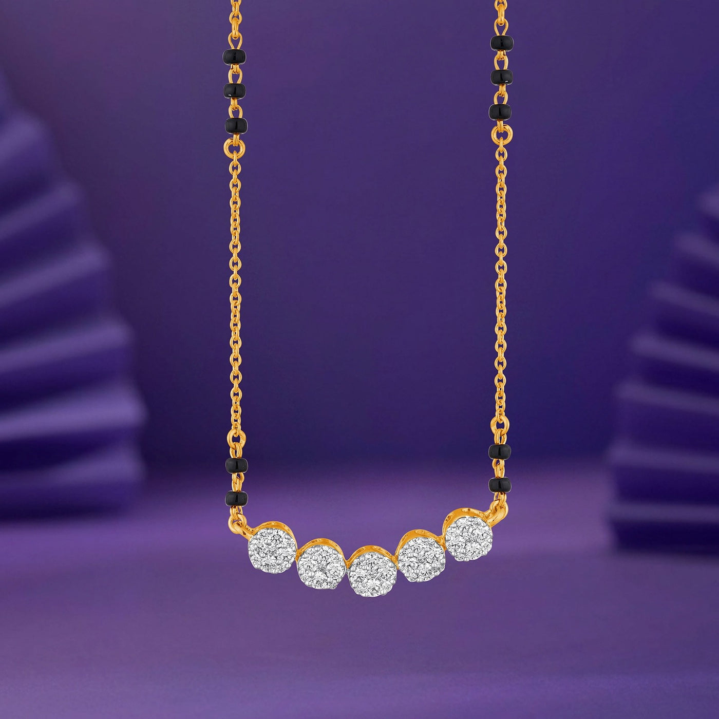 A gold mangalsutra featuring five round, sparkling clear stones set in gold at the center, with small black bead accents along the chain, presented against a purple background.