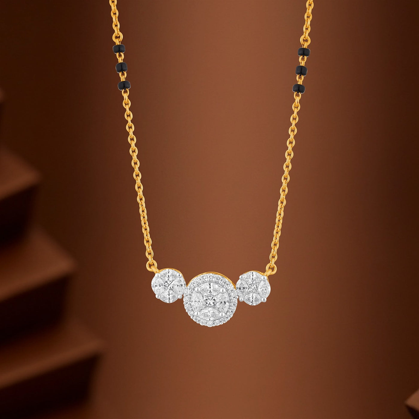 A gold mangalsutra with a pendant featuring three circular diamond clusters in white and yellow gold, set against a warm brown background