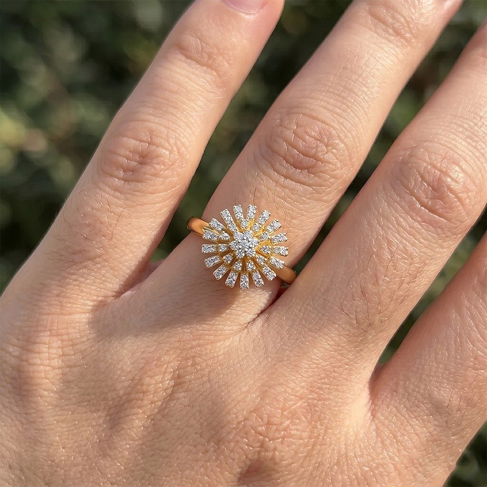 A gold ring with a sunburst cluster of small clear stones, shown worn on a person's finger against a blurred green outdoor background.