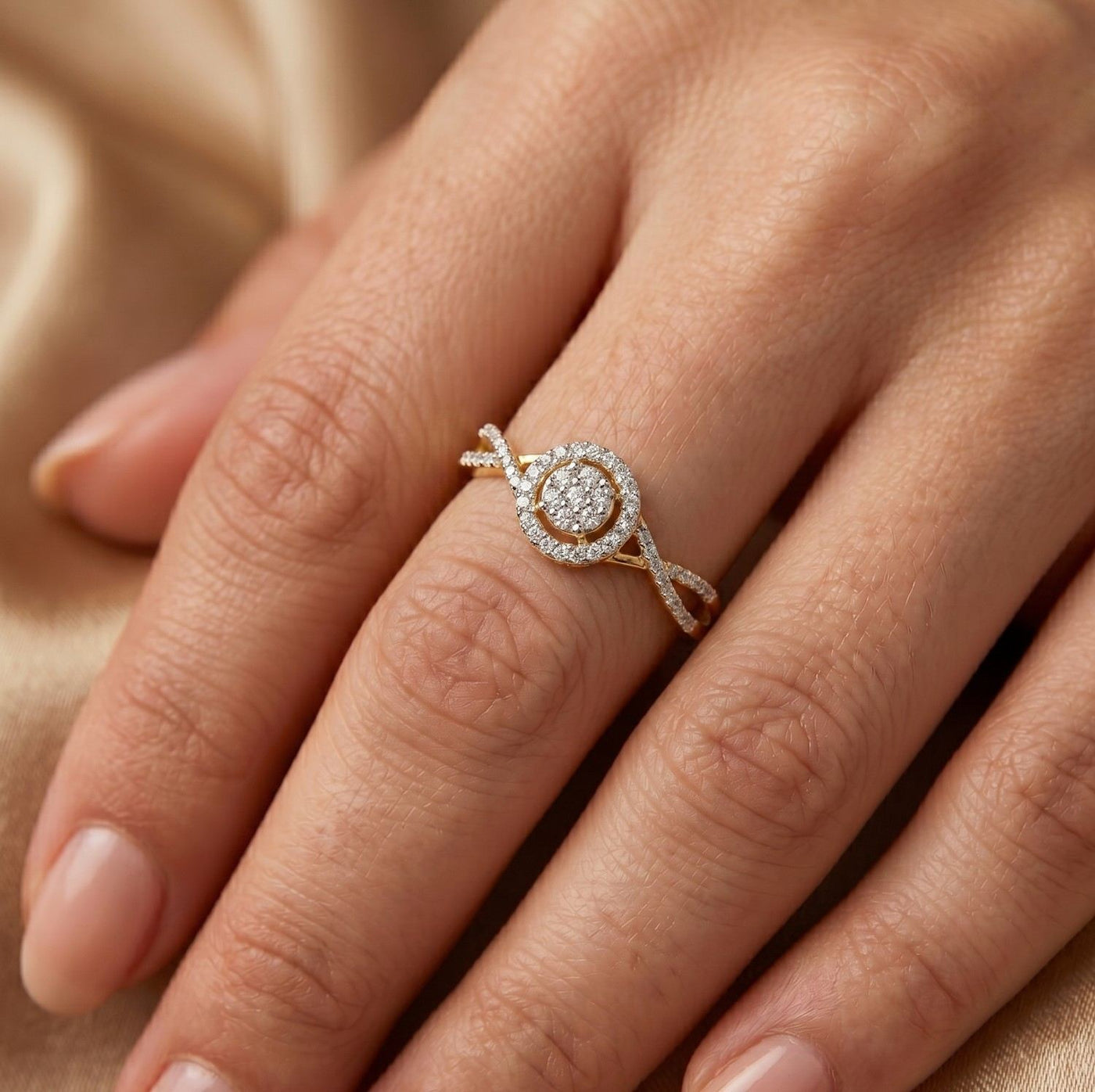A close-up of a hand wearing a gold and diamond halo ring with twisted band details, set against a soft beige fabric background.