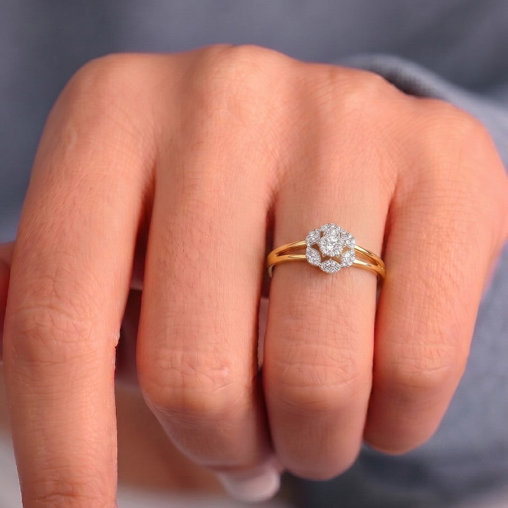 A close-up of a hand wearing a gold ring with a diamond floral design on the ring finger, against a soft gray background.