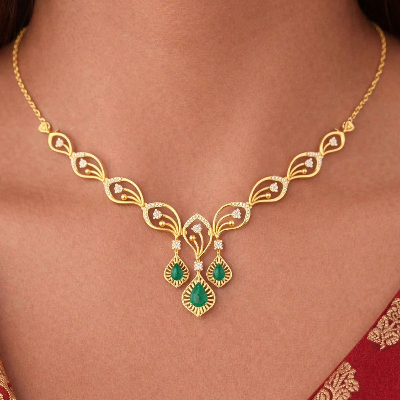 Gold necklace with a symmetrical leaf-shaped design, and two green teardrop gemstone, shown worn on a model's neck.