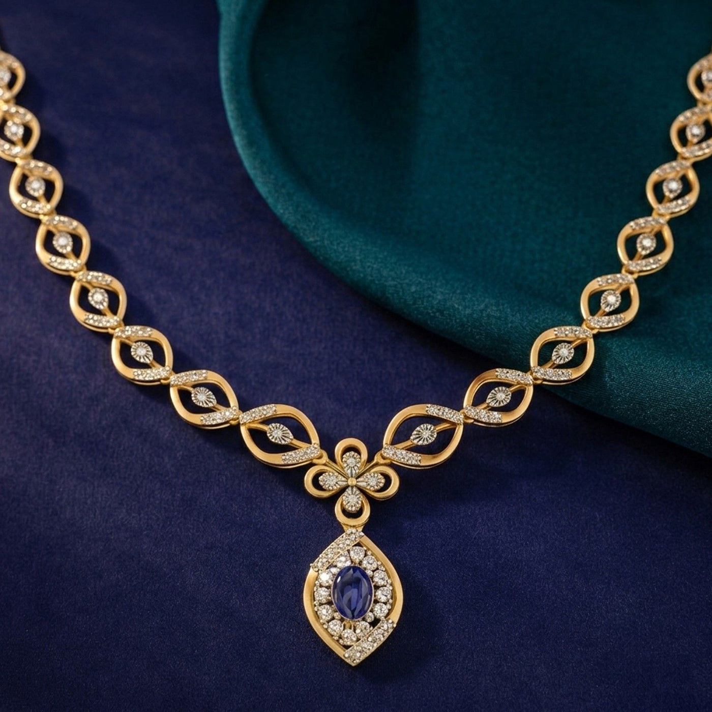 A gold necklace with a teardrop-shaped pendant featuring a sapphire gemstone, displayed on a dark blue fabric background.