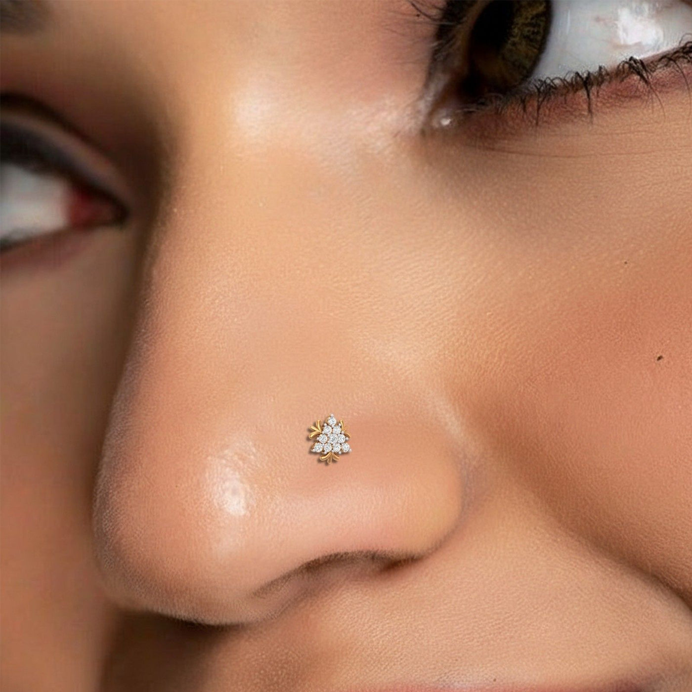A close-up of a person's nose wearing a small gold nose stud pin with clear stones, set against a close-up of the face.