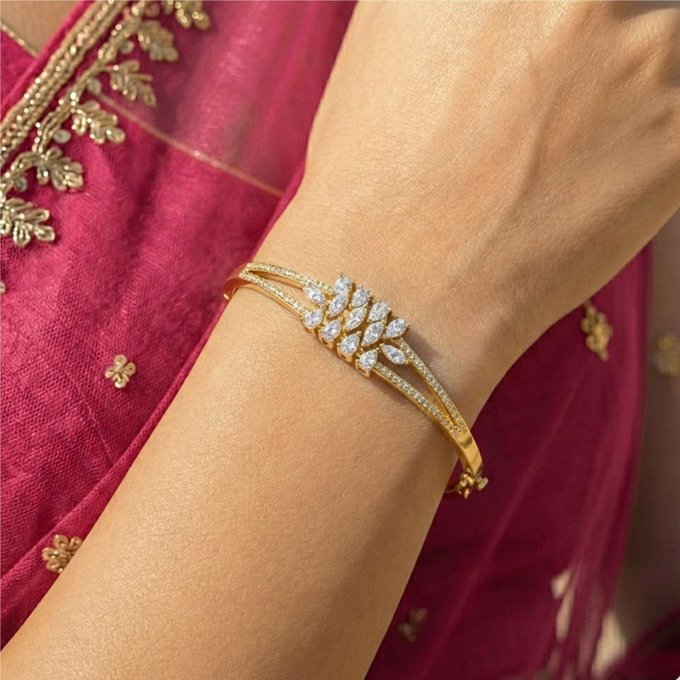 A close-up of a woman's wrist wearing an elegant gold bangle bracelet featuring a triple-band design with a central cluster of marquise-cut white diamonds arranged in a leaf or wheat motif, set against a magenta embroidered silk outfit.