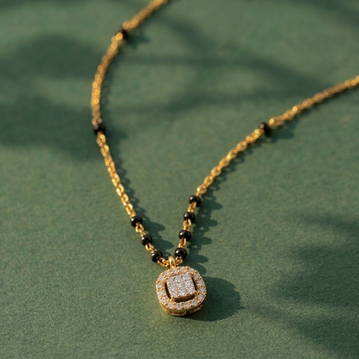 A gold and diamond mangalsutra with small black beads and a square diamond paved with clear stones, displayed on a textured green surface with soft leaf shadows.