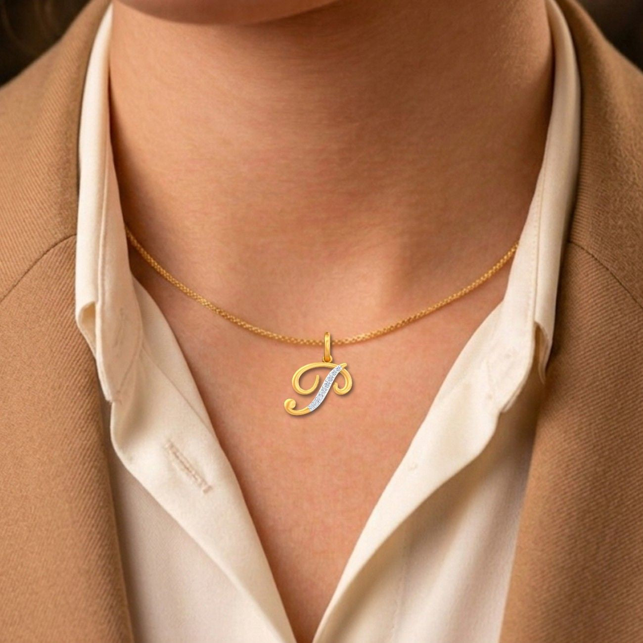 Gold necklace with a 'J' pendant worn by a person in a beige blazer and white shirt.
